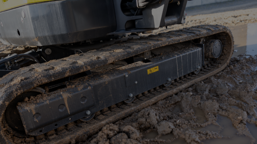 Close-up of a muddy excavator track on wet ground, showing rollers and a yellow warning label on the hull.